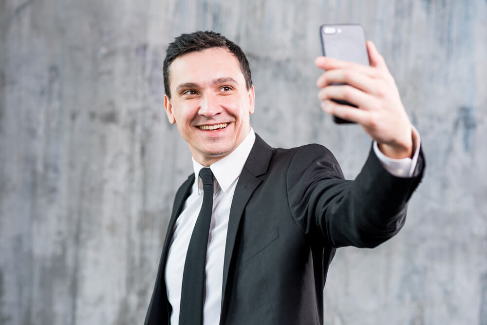 Homem sorrindo segurando o celular com a mão esquerda em posição para retirar uma selfie