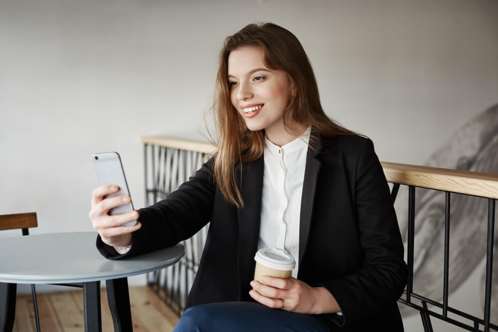 Mulher sorrindo segurando um celular apoiado a mesa com a mão esquerda e um copo na mão direita