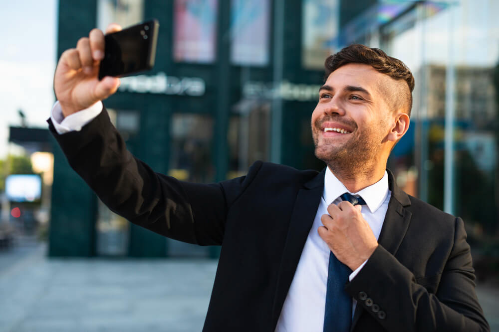 Homem sorrindo segurando o celular com a mão esquerda em posição para retirar uma foto de si