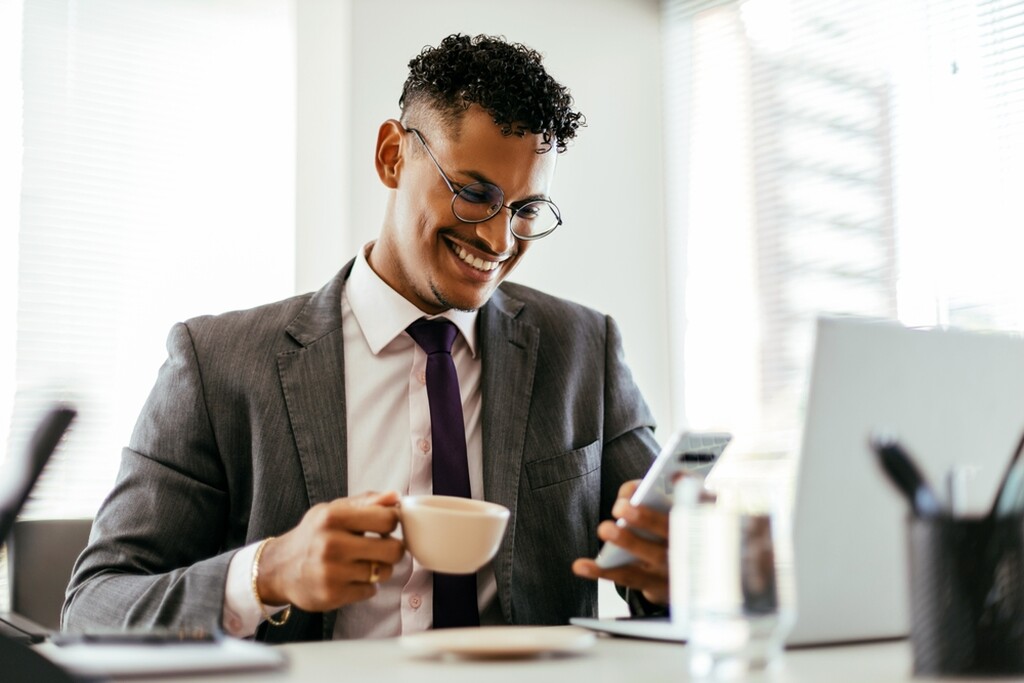 Homem sorrindo e segurando um copo de café, mexendo no celular com um notebook na frente